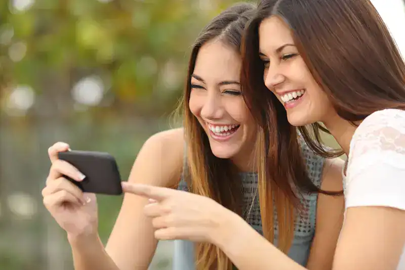 Two young women smiling and sharing a photo on a smartphone outdoors, enjoying a sunny day in a park setting.